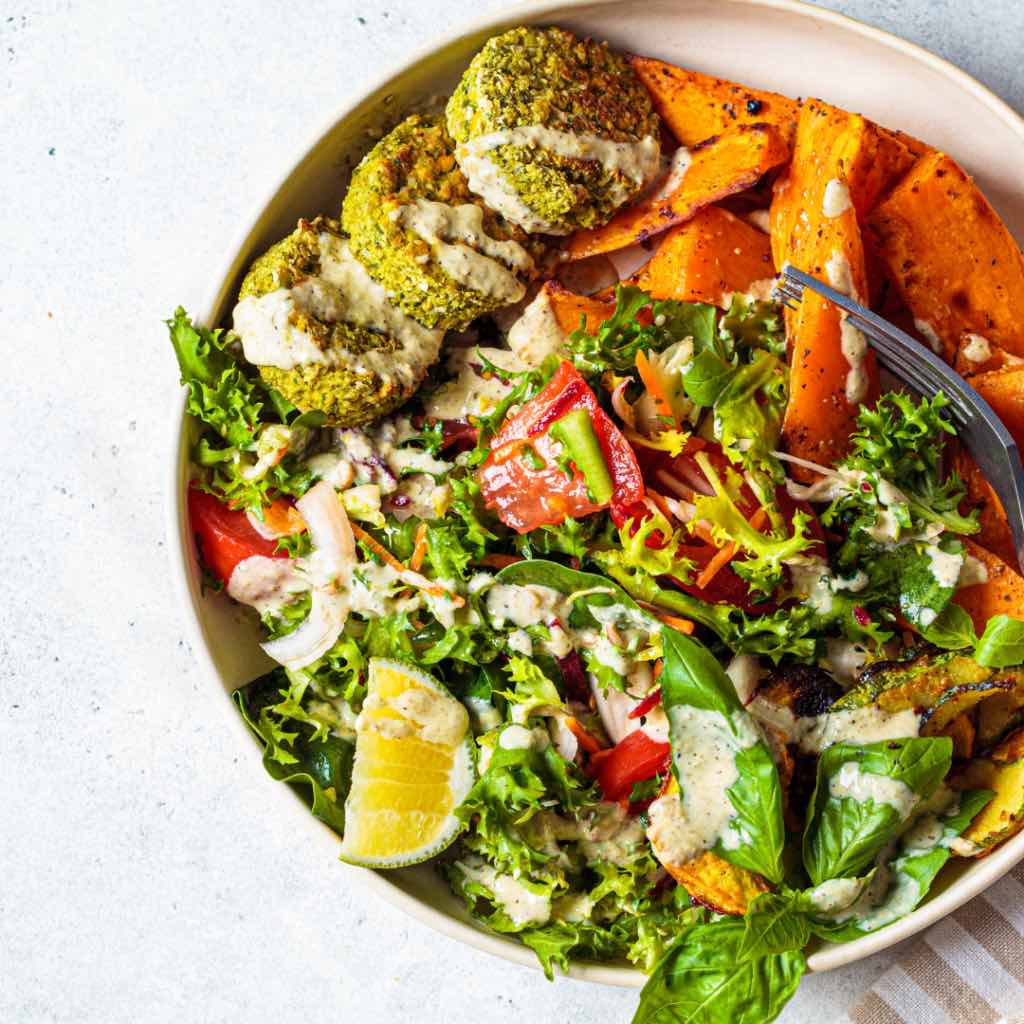 On this picture you see a plate with a salad, green baked falafels and sweet potatoes covered in a tahini dressing.